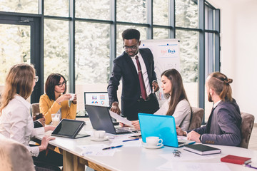 Discussing new business ideas. Cheerful young Afro American Man standing and makes a report near whiteboard while his colleagues sitting at the desk