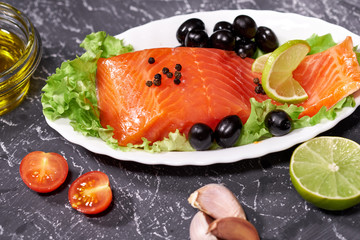 A piece of salmon on a white plate with olives and lemon, on a gray background. Selective focus