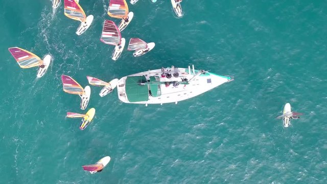 Small white yacht surrounded by a large group of Wind surfers at The Mediterranean Sea - Top down aerial footage.