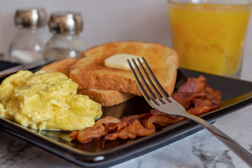 Scrambled eggs, wheat toast with butter, and bacon on square black plate with salt and pepper shakers and orange juice in the background