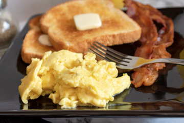 Scrambled eggs, wheat toast with butter and bacon on square plate with fork, up close shot.