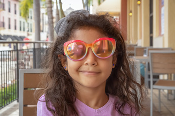 Shabby chic hair topped with a bow compliments the stylish crooked sunglasses of this young girl with a smirk. Having fun sitting outdoors makes for a nicer day.