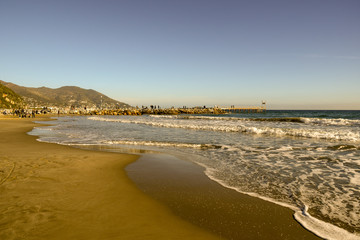 Sandy beach with people walking on a rocky dock in a sunny winter day, Laigueglia, Liguria, Italy
