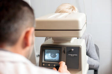 Fototapeta na wymiar Male ophthalmologist performing eye exam using refractometer, looking at patients eye on a screen