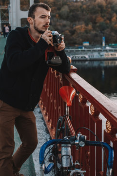 Cyclist photographing with camera while standing with bicycle on bridge in city during sunset