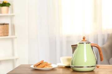 Stylish electrical kettle, cup and plate with cookies on table against blurred room interior. Space for text