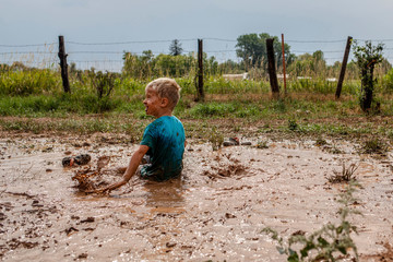 Rear view of playful boy sitting on muddy puddle against sky in yard
