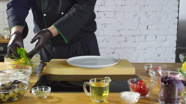 Mid-section tracking shot of unrecognizable male cook in uniform and gloves plating salad. Fresh vegetables, herbs and olive oil are on table
