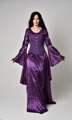 Naklejka premium full length portrait of beautiful girl with long black hair, wearing purple fantasy medieval gown. standing pose on grey studio background.