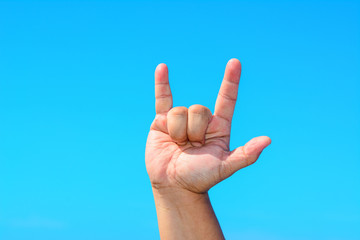 A male hand shows a rock symbol sign with three fingers isolated against blue sky background.