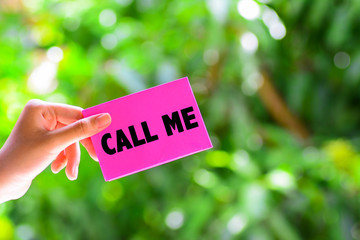 A woman's hand holding a pink color paper with a text 'call me' written on it. Green nature background.