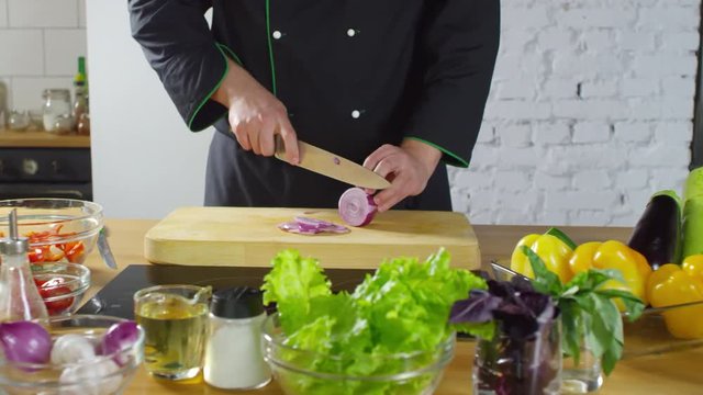 Mid-section tracking shot of unrecognizable male cook in uniform slicing red onion on wooden cutting board while preparing salad. Fresh vegetables and herbs on table
