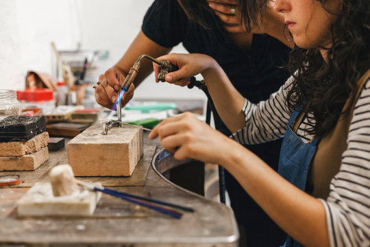 Midsection of teacher assisting female student in using blow torch on ring at workshop