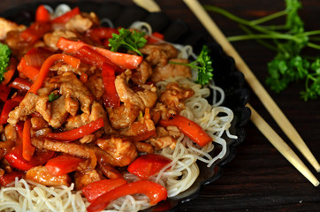 Chinese pork tenderloin with vegetables and noodles on a black saucer on a wooden background