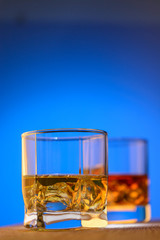 Two glasses of whiskey with pieces of ice on the wood table on a blue background.