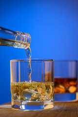 Whiskey from the bottle flowes into the glass with piece of ice on a blue background