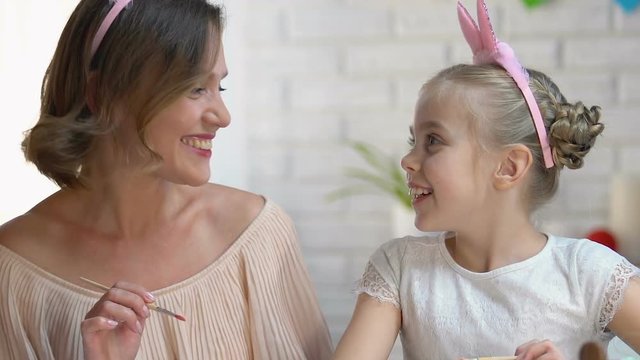Cute daughter kissing mother on cheek decorating Easter eggs, preparing for fest