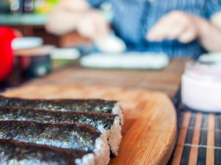Making Sushi Rolls: Rolls Sealing On the Wooden Board before Slicing. Blurred Woman Hands Putting Rice on a Nori Sheet and Kitchenware in Background.