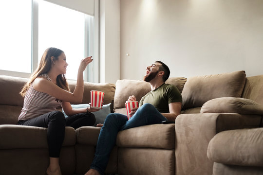 Playful girlfriend throwing popcorn in boyfriend's mouth while sitting on sofa in rental house