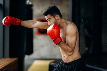 Sportsman with a naked torso and the red boxing gloves on his hands stands hits with right hand in the boxing gym