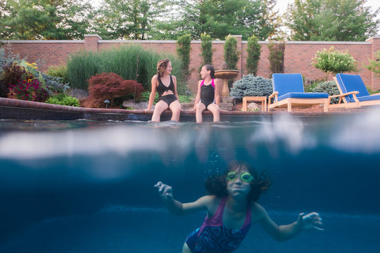 Girl swimming in pool while sisters talking while sitting on poolside - Powered by Adobe