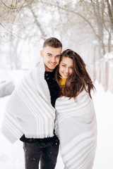 Charming dark-haired girl dressed in a yellow sweater and her boyfriend wrapped together in a gray scarf are standing in a snowy street on a winter day