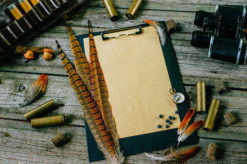 Pheasant feather and cartridges on clipboard with vintage paper for information on old wooden background. Hunting equipment on vintage desk.