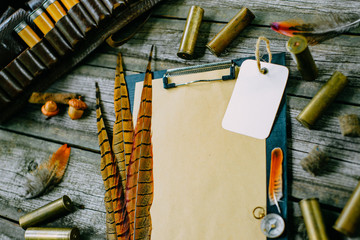 Closeup clipboard with paper and label with pheasant feather and cartridges beside on old wooden...