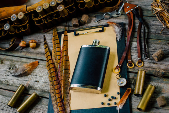 Flask on clipboard and pheasant feather on it on old wooden background. Hunting equipment on vintage desk. Top view