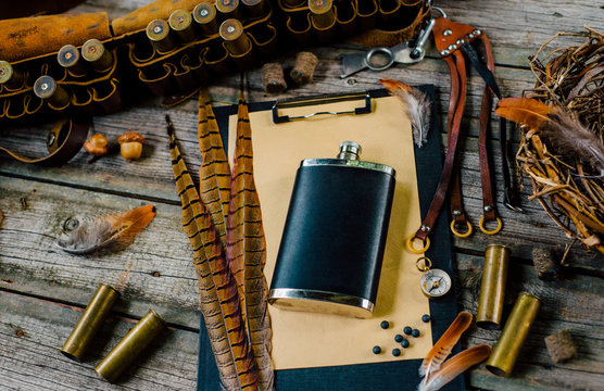 Flask on clipboard and pheasant feather on it on old wooden background. Hunting equipment on vintage desk. Hunting belt with cartridges and tools. Top view