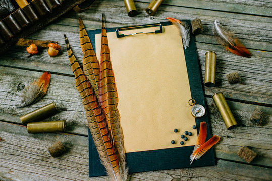 Pheasant feather and cartridges on clipboard with vintage paper for information on old wooden background. Hunting equipment on vintage desk.