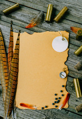 Closeup paper and round label with pheasant feather and cartridges beside on old wooden background....
