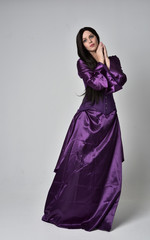 Fototapeta premium full length portrait of beautiful girl with long black hair, wearing purple fantasy medieval gown. standing pose on grey studio background.