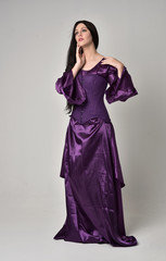 Fototapeta premium full length portrait of beautiful girl with long black hair, wearing purple fantasy medieval gown. standing pose on grey studio background.