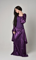 Fototapeta premium full length portrait of beautiful girl with long black hair, wearing purple fantasy medieval gown. standing pose on grey studio background.