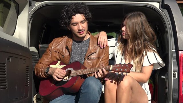 Young man playing guitar for his girlfriend on back of sport utility vehicle