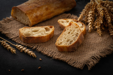 Italian traditional ciabatta bread with sliced ​​pieces and ears of wheat lie on a jute napkin on a table.