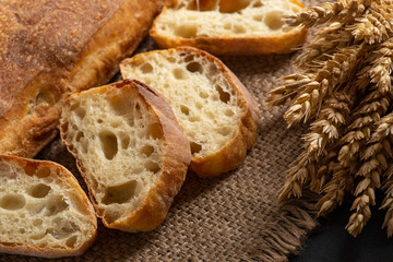 Italian traditional ciabatta bread with sliced ​​pieces and ears of wheat lie on a jute napkin on a table.