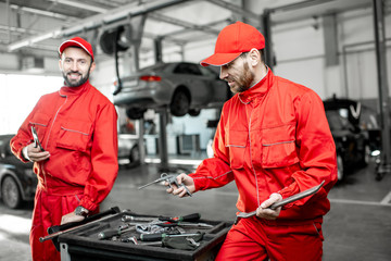 Portrait of a two handsome auto mechanics in red uniform standing with working tools at the car...