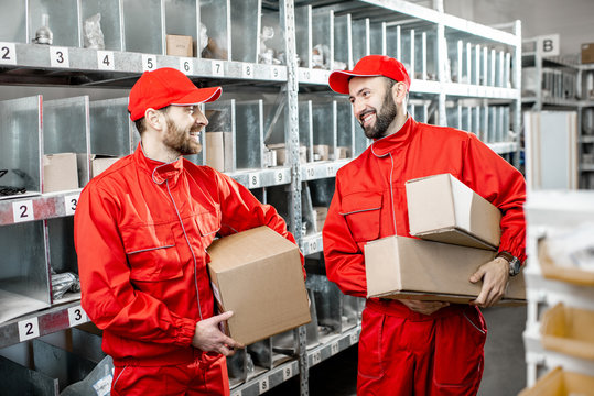 Two handsome warehouse workers in red uniform standing together with boxes in the storage with auto parts or other products