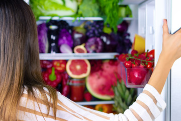 Vegan vegetarian woman female choosing healthy antioxidant colorful veggies, raw juice and fruits for eating after market near opened fridge with: grapefruit, tomatoes, watermelon, pineapple, fig.