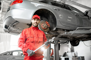 Portrait of a handsome auto mechanic in red uniform standing with mount under the car in the service