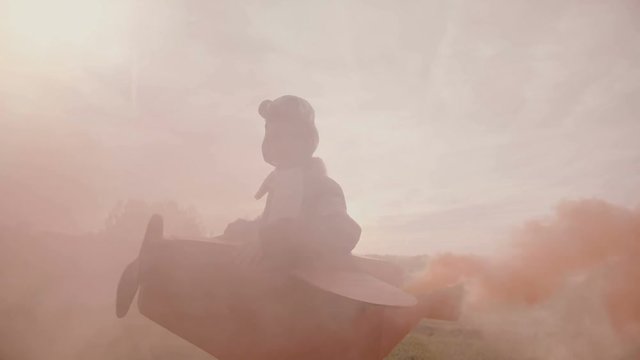 Camera follows little happy smiling girl running in sunset field in fun plane pilot costume with color smoke slow motion