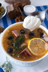 Traditional Russian soup Solyanka cooked with meat, sausages, salted cucumbers on wite bowl. Black bread, lemon, olived and tomatoes on color blue background, homemade food.