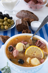 Traditional Russian soup Solyanka cooked with meat, sausages, salted cucumbers on wite bowl. Black bread, lemon, olived and tomatoes on color blue background, homemade food.