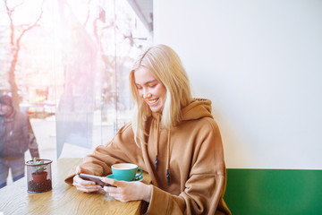 A shot of a beautiful blond student woman lokiing in smart phone, smile at a cafe. - obrazy, fototapety, plakaty