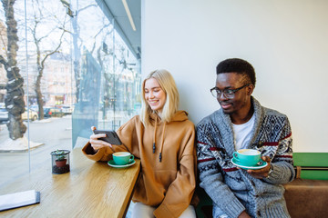 Mixed race couple talking during coffee realx time in cafe. Blond woman and african american man sit together, drinks cappuccino.