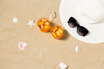 Fototapeta na wymiar vacation, travel and summer concept - two glasses of aperitif cocktails with ice cubes, sun hat, sunglasses and seashells on beach sand
