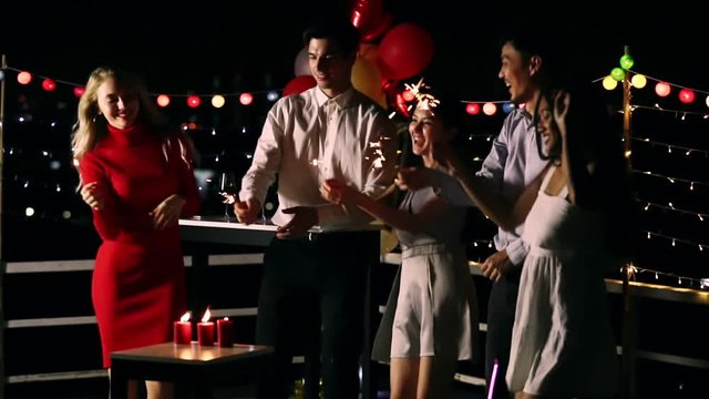Slow motion scene video of Asian and Caucasian people in roof top party, playing sparklers, it's very funny, colorful balloons behind them, concept for New Year party.