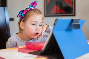 four years old blonde girl with bow headband sitting indoor, with head over hand watching digital tablet with impressed face expression on table